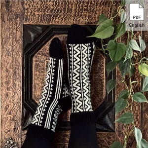 Könnte beinhalten: Ein Paar schwarze Socken mit weißem geometrischen Muster. Die Socken zeigen ein Zickzack-Muster und vertikale Linien. Sie werden vor einem dunklen Holzgrund mit grünen Blättern präsentiert.