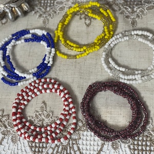 May include: Five beaded waist beads in different colors. The beads are white and blue, white and red, white and maroon, white and yellow, and white and clear.