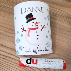 May include: White card with a snowman illustration and the text "Danke Frohe Weihnachten". The snowman is wearing a red scarf and a black hat. The card is surrounded by small blue snowflakes. A red and white chocolate bar with the text "du bist immer so verständnisvoll" is in the foreground.