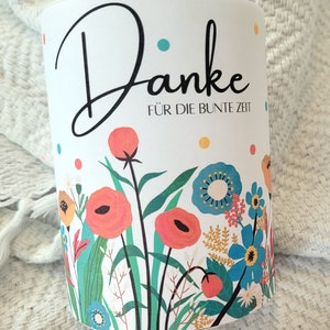 May include: White cylindrical container with a floral design and the text "Danke f&uuml;r die bunte Zeit". The flowers are red, blue, yellow, and green.