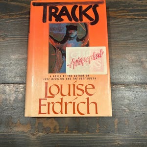 May include: A paperback book titled "Tracks" by Louise Erdrich. The cover features an orange background with a painting of a person in the center. The book is signed "Autographed".