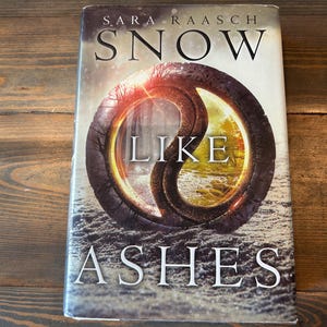 May include: A hardcover book titled "Snow Like Ashes" by Sara Raasch. The cover features a circular design with a yin-yang-like shape in shades of brown, gold, and red. The book is set on a wooden surface.