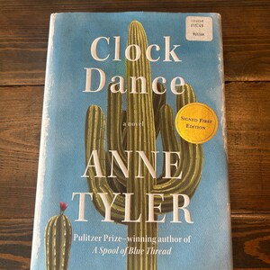 May include: A hardcover book titled "Clock Dance" by Anne Tyler. The cover features a light blue background with a large cactus illustration. A gold sticker reads "Signed First Edition". The book is a novel by the Pulitzer Prize-winning author of "A Spool of Blue Thread".
