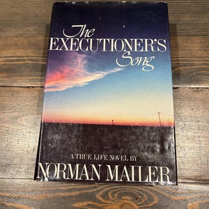 May include: A hardcover book titled "The Executioner's Song" by Norman Mailer. The cover features a sunset landscape with a blue and orange sky. The title is in elegant white script.