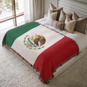 May include: A woven blanket featuring the Mexican flag design. The blanket has a green stripe, a white center with the Mexican coat of arms, and a red stripe. The edges are finished with a fringed trim. The blanket is displayed on a bed.