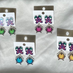 Peut inclure: Boucles d'oreilles pixelisées vertes, roses, bleues et jaunes. Chaque paire présente un motif supérieur et un charm pendant. Les boucles d'oreilles sont présentées sur des cartes blanches, mettant en valeur les motifs colorés inspirés des jeux vidéo.