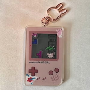 Puede incluir: Llavero rosa Game Girl con un cierre de orejas de conejo en oro rosa. El llavero presenta una pantalla de juego Tetris y el texto "Nantendo GAME GIRL". El diseño incluye un gráfico de gato y piezas de juego pixeladas.