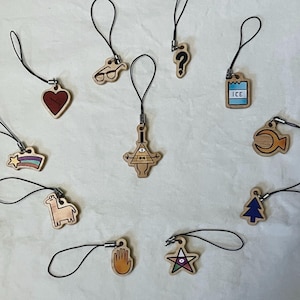 May include: A set of 12 wooden charms featuring characters and symbols from the animated series Gravity Falls. The charms are attached to black string and are perfect for adding a touch of whimsy to your keys, bags, or phone.