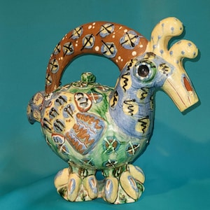 May include: A whimsical ceramic teapot in the shape of a rooster. The teapot features a brown handle with white and blue accents, a blue head, and a body with green, yellow, and blue patterns. The rooster has a yellow comb and feet.