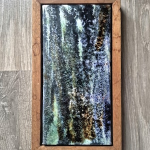 May include: Rectangular ceramic tile with a textured, abstract design in shades of blue, green, and brown, set within a wooden frame. The artwork features vertical streaks and a mottled appearance, creating a unique visual texture.