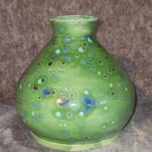 May include: A green ceramic vase with a bulbous body and a narrow neck. The vase is speckled with blue and brown spots, creating a unique, textured appearance. The vase is approximately 7 inches tall.