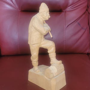 May include: A carved wooden figure of a man wearing a plaid shirt, a knitted hat, and boots, holding a wooden tool. The figure stands on a wooden base with a log. The carving is a light brown colour.