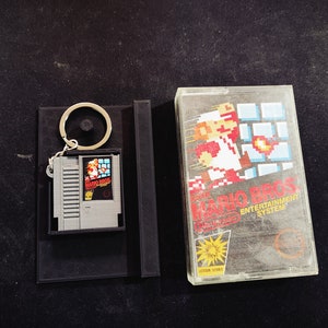 May include: A gray Mario Bros. video game cartridge keychain with a black holder and a clear plastic game case. The game cartridge features the Mario Bros. logo and character art. The case has the game title and logo.