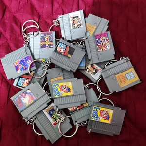 May include: A collection of gray NES cartridge keychains with various game labels, including Super Mario Bros. 3 and The Legend of Zelda. Each keychain has a silver metal ring. The keychains are arranged on a burgundy surface.
