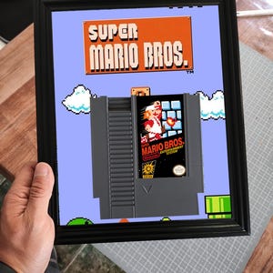 Pode incluir: Obra de arte emoldurada de Super Mario Bros. A imagem apresenta o logotipo do jogo em laranja e branco, com um fundo pixelizado e um cartucho de jogo cinza com o rótulo do jogo visível.