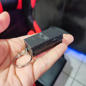 Może przedstawiać: Czarny plastikowy brelok w kształcie konsoli PlayStation 2. Brelok ma srebrny metalowy pierścień.