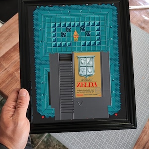 Puede incluir: Obra de arte enmarcada con un mapa pixelado en verde azulado y azul, con un cartucho de videojuego dorado y gris. El cartucho dice "The Legend of Zelda" e incluye un logotipo de Nintendo Entertainment System. El marco es negro.