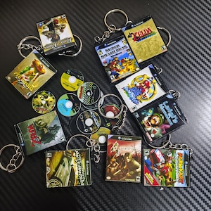Może przedstawiać: Kolekcja miniaturowych breloków do kluczy w kształcie pudełek na gry Nintendo GameCube z tytułami takimi jak Super Smash Bros. Melee, The Legend of Zelda, Luigi's Mansion, Metroid Prime, Resident Evil 4 i Mario Kart: Double Dash oraz małymi okrągłymi płytami z grami.