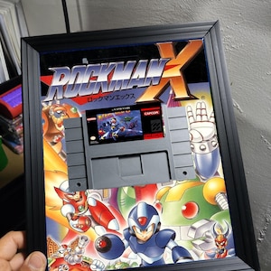 May include: Framed Rockman X Super Nintendo game cartridge. The artwork features a vibrant illustration of characters in action, with the game title prominently displayed. The cartridge is gray and set against a colorful background, all within a black frame.