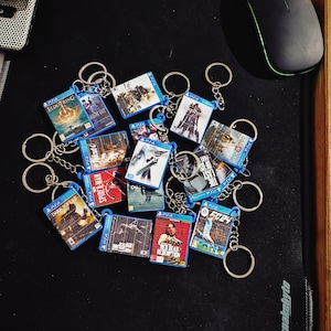 Może przedstawiać: Kolekcja breloków do kluczy z popularnymi grami wideo, takimi jak "Dark Souls", "Spider-Man", "Red Dead Redemption", "The Last of Us" i "Elden Ring". Breloki są wykonane z plastiku i mają kolorowe grafiki gier.