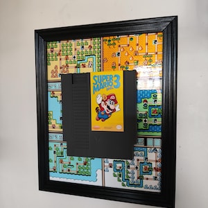 May include: Framed Super Mario 3 Bros. video game cartridge. The black cartridge is centered on a colorful map background, all within a black frame. The game's label features Mario in mid-air.