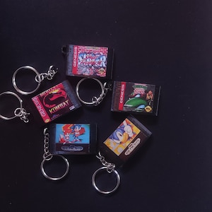 Peut inclure: Cinq porte-clés de cartouche de jeu Sega Genesis. Les porte-clés représentent les jeux : Mortal Kombat, Super Street Fighter II, Teenage Mutant Ninja Turtles IV : Turtles in Time, Sonic the Hedgehog 2 et Sonic the Hedgehog 3. Chaque porte-clés est noir avec un anneau de clé argenté.