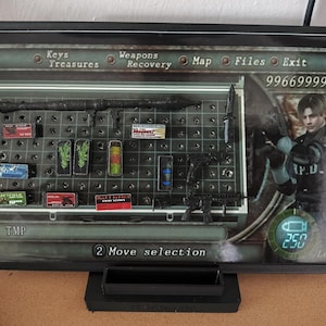 May include: A framed display featuring a video game inventory screen. The screen shows weapons, ammunition, and other items. The character Leon is visible on the right. The text includes "Keys", "Treasures", "Weapons", "Recovery", "Map", "Files", "Exit", "TMP", and "2 Move selection".