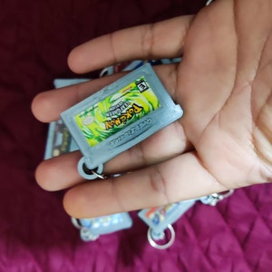 May include: A close-up of a gray Game Boy Advance game cartridge keychain with a green and white Pokemon Leaf Green label. The label features the Pokemon logo and the text "Pokemon Leaf Green Version".