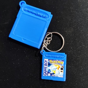 May include: Blue plastic keychain shaped like a Game Boy cartridge with the text "GAME BOY" and "Pokémon" on the front. The cartridge features a blue and white image of a Pokémon character and the text "Blue Version".