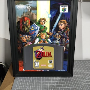 Może przedstawiać: Oprawiona grafika z kartridżem do gry Nintendo 64 "The Legend of Zelda: Ocarina of Time". Grafika zawiera okładkę gry i czarną ramę. Kartridż do gry jest szary i żółty.