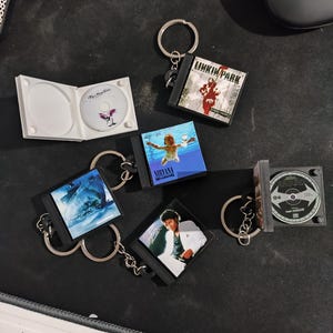 Puede incluir: Llaveros con estuches de CD en miniatura con portadas de álbumes de varios artistas, incluidos Nirvana y Linkin Park. Los llaveros son negros y plateados, con un llavero plateado. Un llavero tiene una caja de CD abierta.