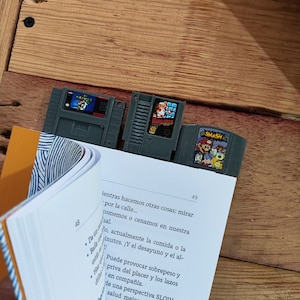 May include: Three gray video game cartridge bookmarks are placed on the pages of a book. The bookmarks feature the game titles "Mario Bros" and "Smash". The book is open to pages 48 and 49.