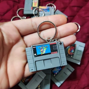 May include: A collection of Super Nintendo Entertainment System (SNES) game cartridge keychains. The keychains are gray and feature various game titles, including "Super Mario World".