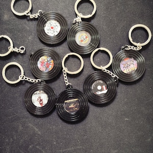 May include: Seven black vinyl record keychains with silver key rings. Each keychain features a different band logo, including KISS, Guns N' Roses, and Iron Maiden.