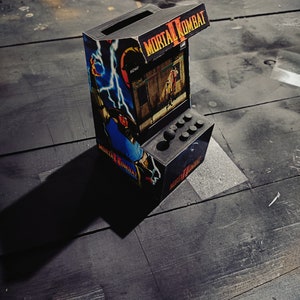 Puede incluir: Un gabinete de arcade en miniatura con un exterior negro y un diseño colorido de Mortal Kombat II. El gabinete tiene una pantalla pequeña, botones y una palanca de mando.