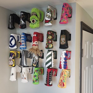 May include: A wall display of colorful golf headcovers with various designs and logos. The headcovers are hung on a white wall with black hooks. Some of the headcovers feature text such as "Pine Tree Golf Club", "Par 3", "Medford Lakes", and "Cameron".