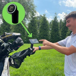 May include: A black golf cart phone holder with a rectangular screen is pointed at by a person. The image also shows a black golf club head cover and a black golf club accessory with the text "LUNA'S" on it.