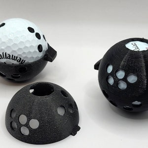 May include: Three black 3D printed golf ball holders. One holder has a white golf ball inside with the word "Callaway" printed on it. Another holder has a white golf ball inside with the word "Quik" printed on it. The third holder is empty.