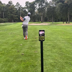 Puede incluir: Un golfista balancea un palo de golf en un campo de golf verde. Un trípode negro con un teléfono inteligente conectado a él está grabando al golfista.