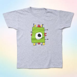Könnte beinhalten: Hellgraues T-Shirt mit einem grünen Monster-Motiv. Das Monster hat ein großes weißes Auge, rote Stacheln, gelbe Füße und schwarze Arme. Das Motiv ist ein grünes Quadrat. Das T-Shirt ist aus weichem Material.