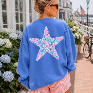 May include: A blue sweatshirt with a large starfish design on the back. The starfish is filled with a floral and seashell pattern in pink, blue, and white. The person is wearing pink and white checkered shorts.