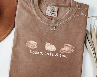 May include: A brown t-shirt with the words "books, cats & tea" printed below a stack of books, a cat, and a teacup. The shirt has a crew neck and short sleeves. A silver necklace with a small pendant is draped across the shirt.