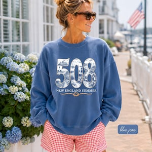 Puede incluir: Una sudadera azul de cuello redondo con los grandes números blancos 508 rellenos de ilustraciones costeras en azul y blanco de un faro, un ferry y escenas de playa. Debajo de los números aparece el texto NEW ENGLAND SUMMER y un nudo de cuerda.