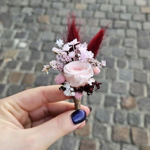 Op de afbeelding: Een kleine, handgemaakte bloemenboutonnière met een lichtroze roos, bordeauxrode accenten en kleine witte en roze bloemen. De compositie bevat bordeauxrode, veervormige pluimen en een met touw omwikkelde stengel. Perfect voor bruiloften.