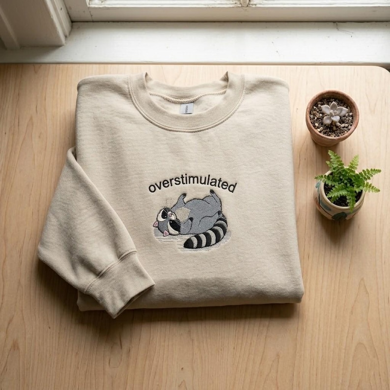 Puede incluir: Sudadera beige con la palabra "overstimulated" bordada sobre un mapache de dibujos animados tumbado boca arriba. El mapache es gris con una cola a rayas blancas y negras. Una peque&ntilde;a suculenta y un helecho en maceta est&aacute;n junto a la sudadera.