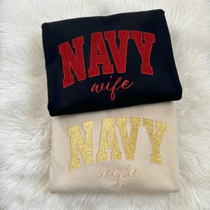 May include: Two sweatshirts, one black and one beige, are stacked. The black sweatshirt has the word "NAVY" in red glitter and "wife" in cursive. The beige sweatshirt has "NAVY" in gold glitter and "wife" in cursive. Both are folded.