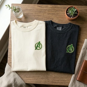 May include: Two t-shirts, one cream-colored and one black, are displayed on a wooden surface. Each shirt features a green embroidered Avengers logo on the chest. The shirts are neatly folded, showcasing their simple design.