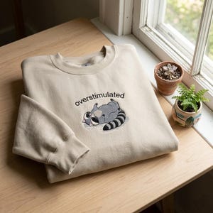 Puede incluir: Sudadera beige con la palabra "overstimulated" bordada sobre un mapache de dibujos animados. El mapache es gris, negro y blanco, tumbado boca arriba. La sudadera est&aacute; doblada sobre una superficie de madera cerca de una ventana.