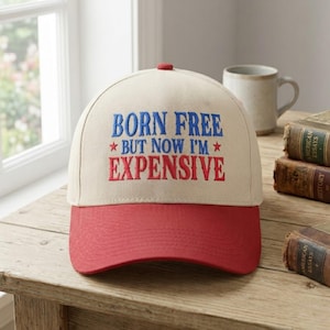 May include: A baseball cap with a cream-colored crown and a red brim. The cap features the embroidered text "BORN FREE * BUT NOW I'M * EXPENSIVE" in blue and red. A red button is on top of the cap.