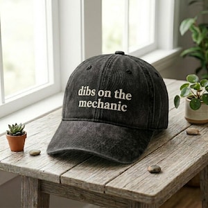 May include: A black baseball cap with the phrase "dibs on the mechanic" embroidered in white. The cap is made of a worn-looking fabric and sits on a weathered wooden table near a window.